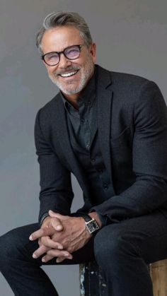 an older man in a suit and glasses sitting on a stool smiling at the camera