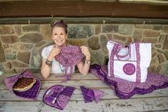 a woman sitting at a picnic table with purple cloths on it and two bags next to her