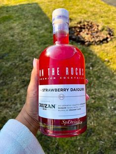 a person holding up a bottle of strawberry daiquit in the grass near a park