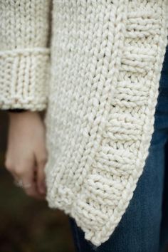 a close up of a person wearing a white knitted sweater and jeans with their hands in the pockets