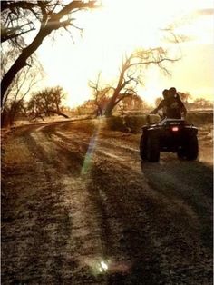 two people riding on four wheelers down a dirt road at sundown in the desert