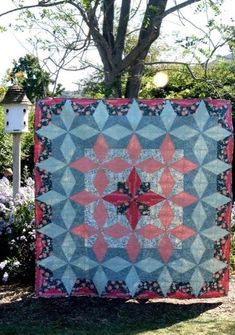 a quilt is hanging on the side of a tree in front of some bushes and flowers