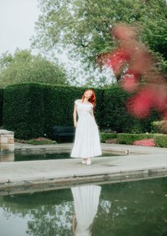 a woman in a white dress is standing by a pond and looking at the camera