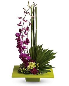 an arrangement of purple flowers and green plants in a square vase on top of a table