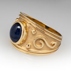 This artisanal ring is centered with one (1) oval cabochon cut natural sapphire set into a bezel setting. The ring features a high polished, raised scroll and beaded details atop a matted background. The ring measures 13.5mm at the top, rises 5.6mm above the finger, tapering to 3.7mm wide and 0.8mm thick at the base of the shank. It is currently a size 7. Formal 14k Gold Sapphire Ring With Oval Cabochon, Heirloom Style Oval Cabochon Sapphire Ring, Elegant Hand Forged Oval Moonstone Ring, Elegant Hand Forged Moonstone Ring, Heirloom Style Sapphire Ring With Oval Cabochon, Elegant Hand Forged Sapphire Ring For Anniversary, Hand Forged Oval Sapphire Ring For Anniversary, Heirloom Sapphire Ring With Oval Cabochon, Antique Cabochon Sapphire Ring For Formal Events