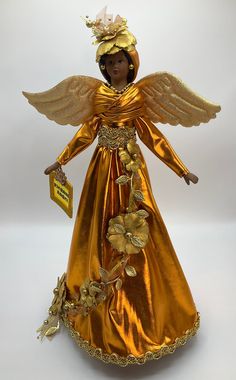 an angel figurine with gold wings and flowers on it's head, holding a tag