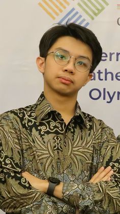 a young man wearing glasses standing in front of a sign with his arms crossed and looking at the camera