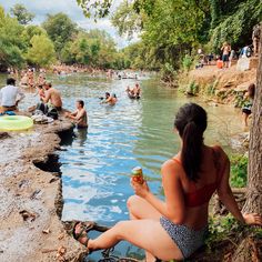 a woman sitting on the edge of a body of water with people swimming in it