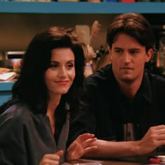 a man and woman sitting at a table in front of a book shelf with books on it