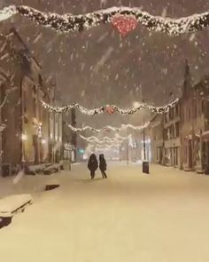 two people walking down a snowy street with lights strung over the buildings and on the sidewalk