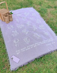 a blanket with flowers on it laying in the grass next to a basket and cell phone