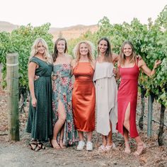 four women in dresses standing next to each other near some trees and bushes with their arms around one another