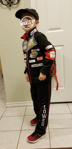 a young boy wearing a racing suit and hat, standing in front of a door