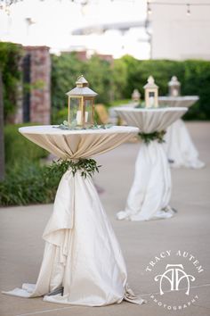 two white tables topped with gold lanterns and draped tablecloths are decorated with greenery