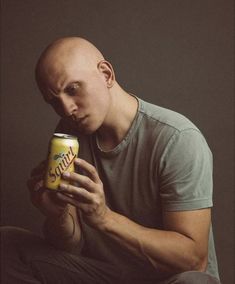 a bald man sitting down holding a can of soda in his right hand and looking at the camera