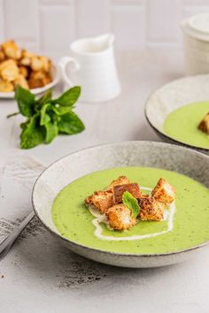 two bowls filled with green soup and croutons on top of a white table