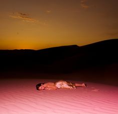 a woman laying on top of a sandy beach under a pink sky at sunset in the desert