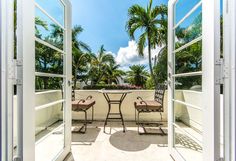 an open door leading to a balcony with two chairs and tables on it, surrounded by palm trees