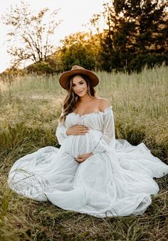 a pregnant woman wearing a white dress and hat sitting on the ground in tall grass