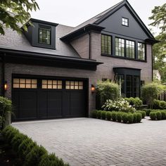 a large brick driveway leading to a house with black garage doors and two story windows