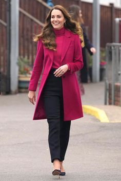 a woman walking down the street in a red coat and black pants with her hand on her hip