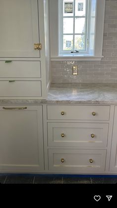 a white kitchen with marble counter tops and gold pulls on the cabinets in front of a window