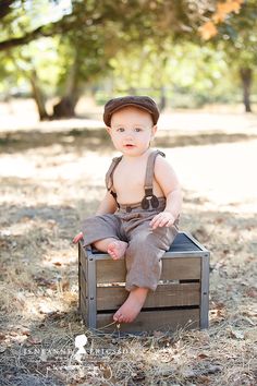 a baby boy sitting on top of a crate in the grass wearing a hat and overalls