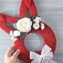 a person holding a red knitted wreath with white flowers and greenery on it