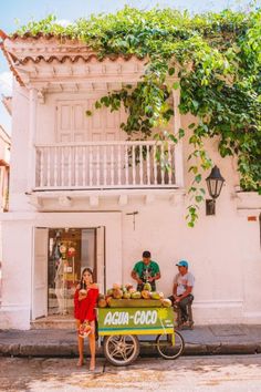 two people standing in front of a building with a fruit cart on the street next to it