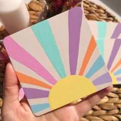 a hand holding four colorful coasters in front of a basket with flowers and milk