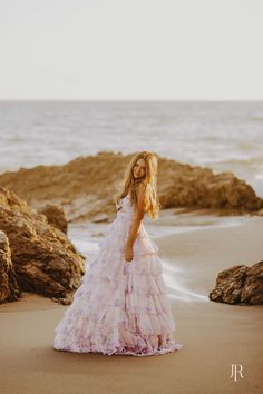 a woman in a long dress standing on the beach next to some rocks and water