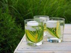 three glasses filled with water and lemon slices on top of a wooden table next to green grass