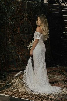 a woman standing in front of a rug wearing an off the shoulder white wedding dress