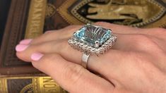 This marvelous ring is centered with one (1) emerald cut natural aquamarine set into a four-prong setting. The center stone is bordered with twenty-six (26), prong set, round brilliant cut diamonds. The ring measures 23.1mm at the top, rises 12.6mm above the finger, tapering to 3.4mm wide and 0.9mm thick at the base of the shank. This ring is currently a size 6.25. Aquamarine Cocktail Ring, Natural Aquamarine, Round Brilliant Cut Diamond, Cocktail Ring, Brilliant Cut Diamond, Cocktail Rings, Emerald Cut, Round Brilliant, Prong Setting