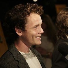 a man with curly hair is smiling and talking into a microphone while another person looks on