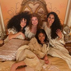 four women in white robes are sitting on a bed with gold sheets and pillows, posing for the camera