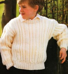 a young boy wearing a white sweater standing in front of a fence with his hands on his hips