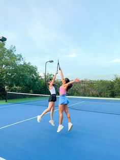 two women are playing tennis on a blue court with trees in the background and one woman is reaching up to hit the ball