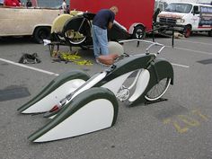 a man standing next to a green and white motorcycle in a parking lot with other cars