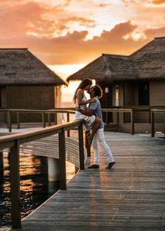 a man and woman kissing on a dock at sunset with thatched huts in the background