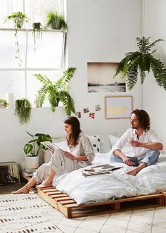 two people sitting on a bed with white sheets and pillows, one reading a book