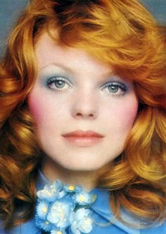 a woman with red hair wearing a blue shirt and flower collared necklace, looking directly into the camera