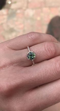 a person's hand holding a ring with a green stone in the center and diamond band around it