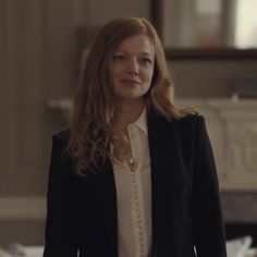 a woman with long hair wearing a black jacket and white shirt standing in front of a bed