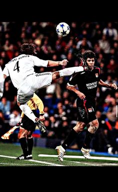 two men are playing soccer in front of an audience at a sporting event and one is jumping up to head the ball