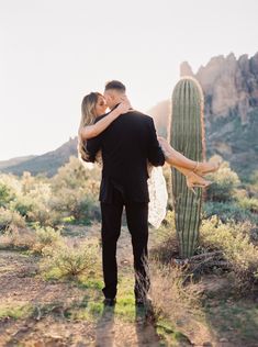 a man holding a woman in his arms next to a cactus with mountains in the background