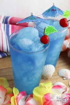 two blue cocktails sitting on top of a wooden table next to shells and flowers