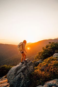 a woman standing on top of a large rock with the sun setting in the background