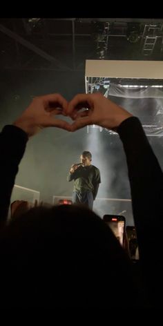 someone making a heart with their hands in front of a concert stage while another person holds his hand up to the camera