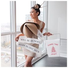 a woman in a dress and hat holding two bags with words on them, while standing next to a window